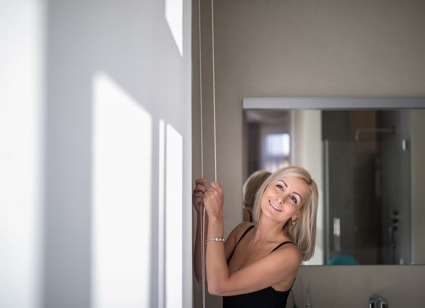 Pretty, young woman lowering the interior shades/blinds in her modern interior apartment